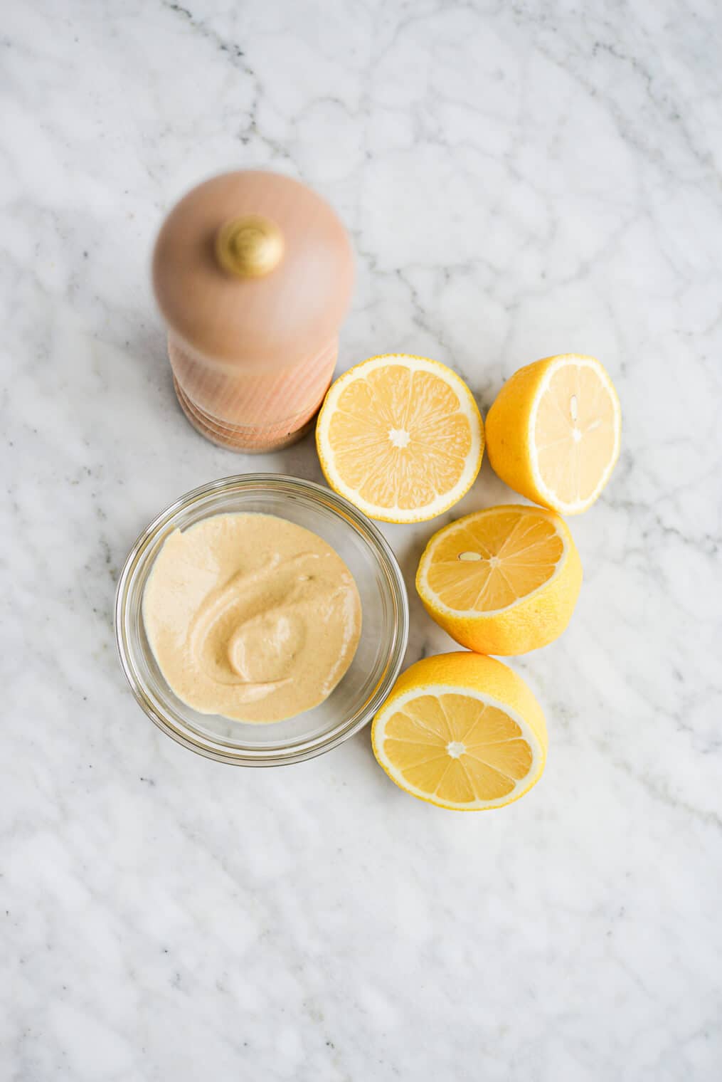 a small bowl of dijon mustard, two halved lemons, and a pepper grinder sitting on a marble surface