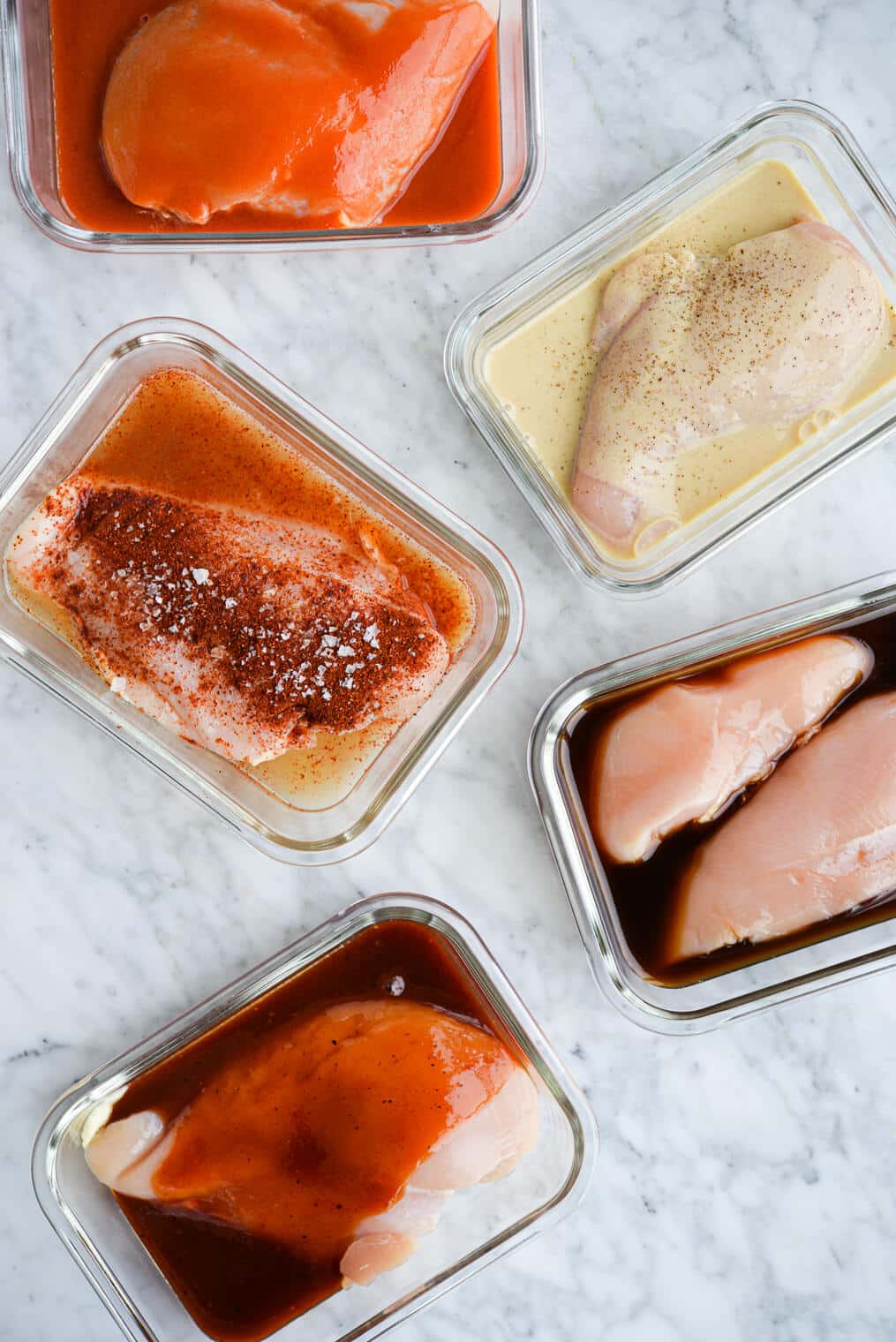 5 glass containers filled with raw chicken and 5 different chicken marinades (lemon dijon, teriyaki, buffalo, BBQ, and taco) sitting on a marble surface