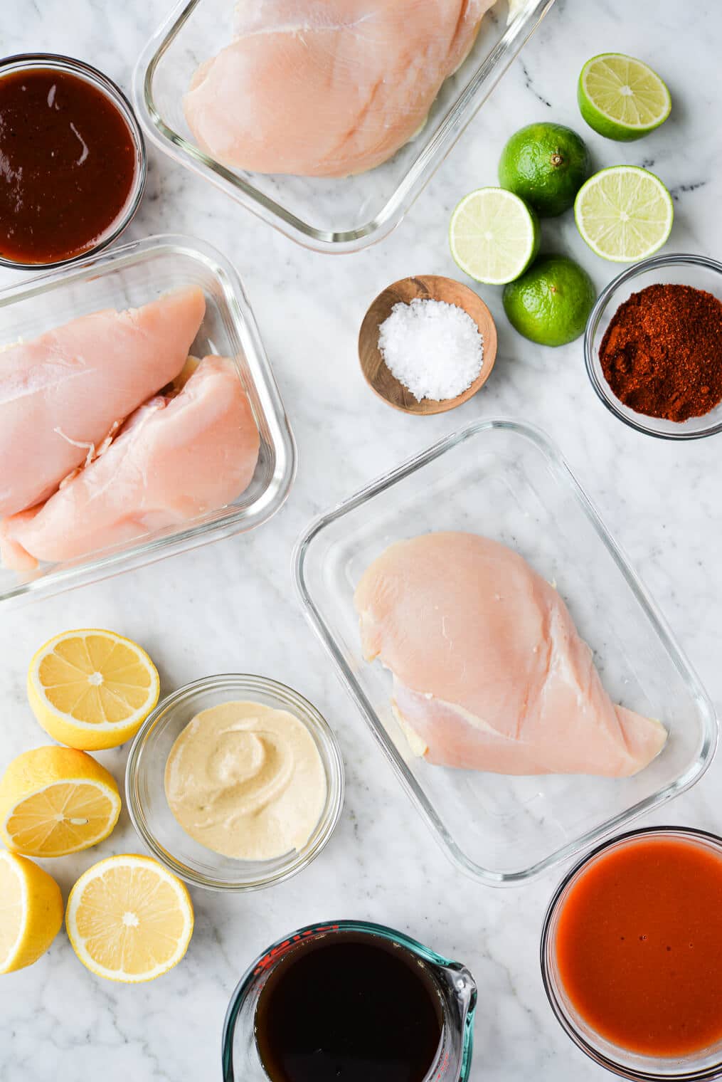 three meal prep containers with raw chicken breasts, and the ingredients for several different chicken marinades sitting on a marble surface