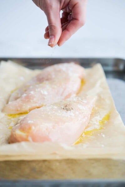 a woman's hand sprinkling ground pepper on raw chicken breasts that are sitting on a parchment paper lined sheet pan and coated in olive oil and sea salt