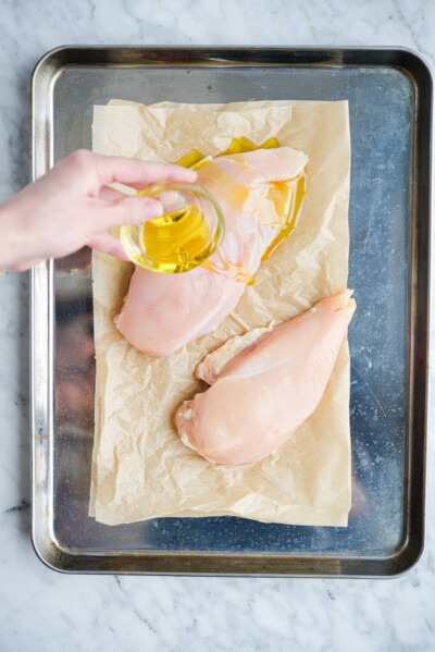 a woman's hand pouring a small bowl of olive oil over a chicken breast sitting on a parchment paper lined sheet pan on a marble surface