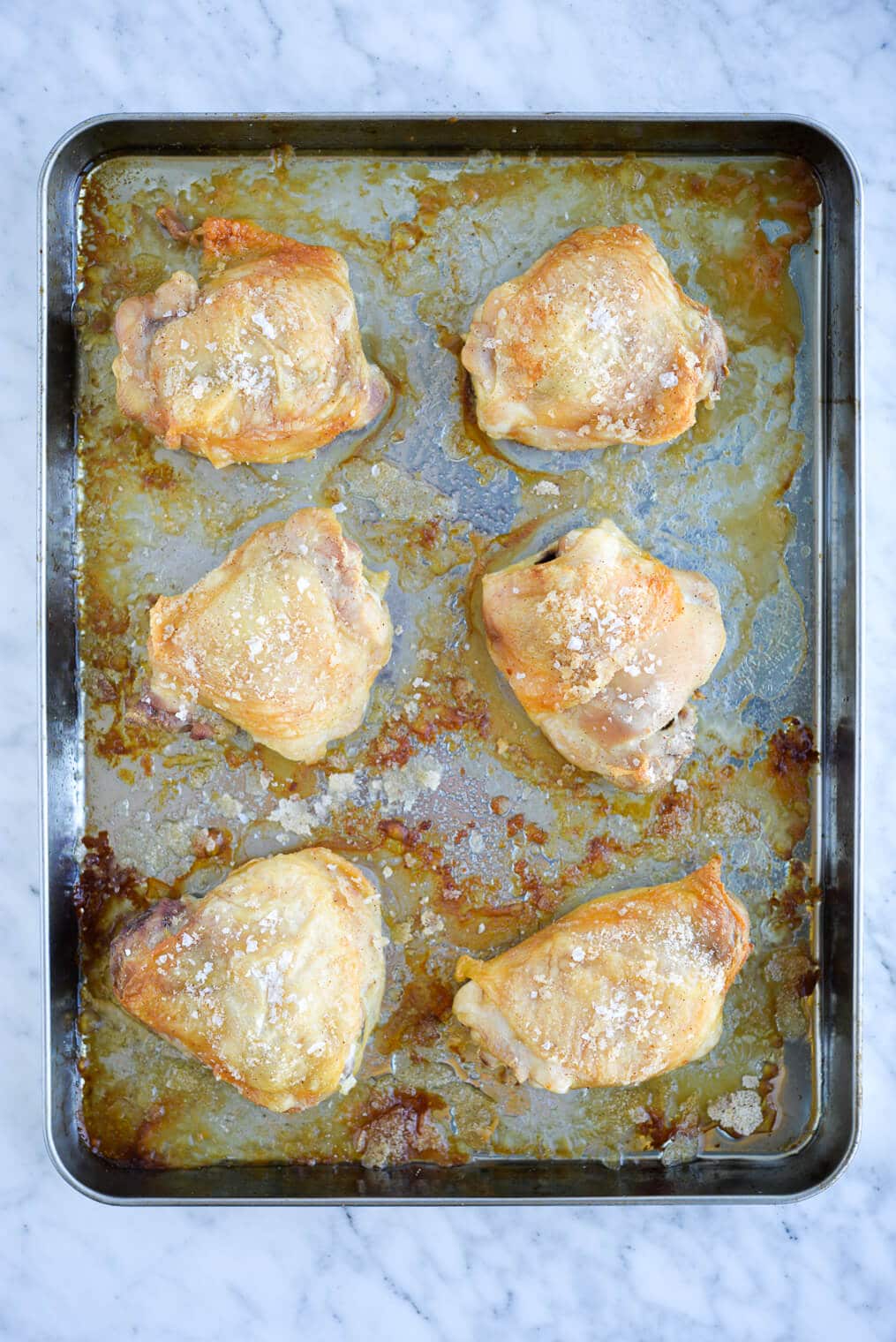 golden brown oven baked crispy chicken thighs on a stainless rimmed sheet pan sitting on a marble surface