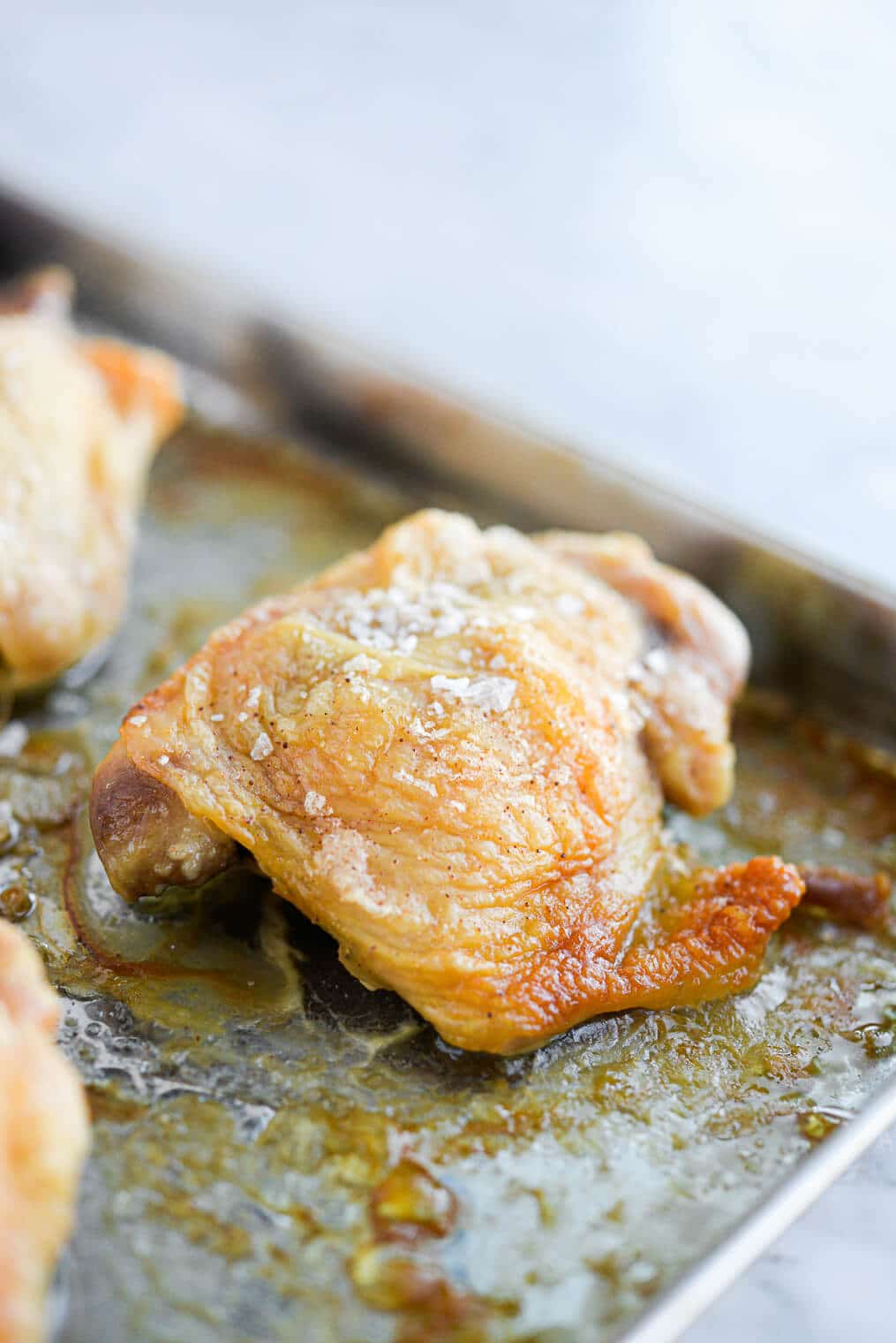 a golden brown oven baked crispy chicken thigh on a stainless rimmed sheet pan sitting on a marble surface