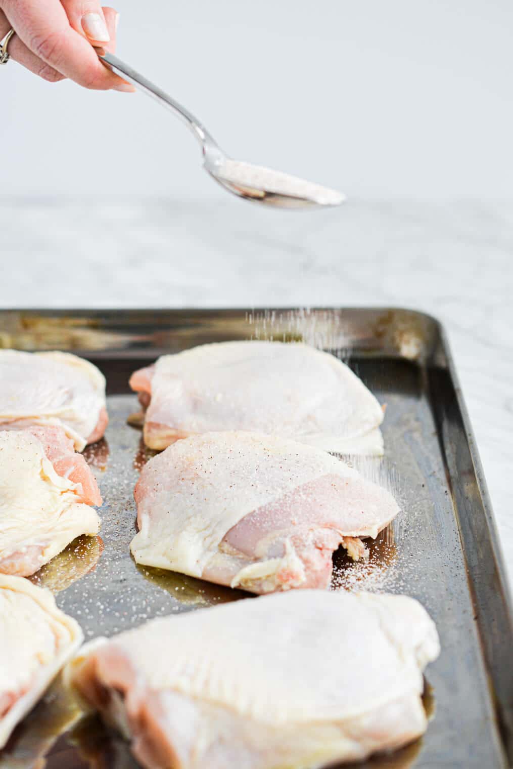 a woman sprinkling sea salt over bone-in, skin-on chicken thighs laying on a rimmed baking sheet