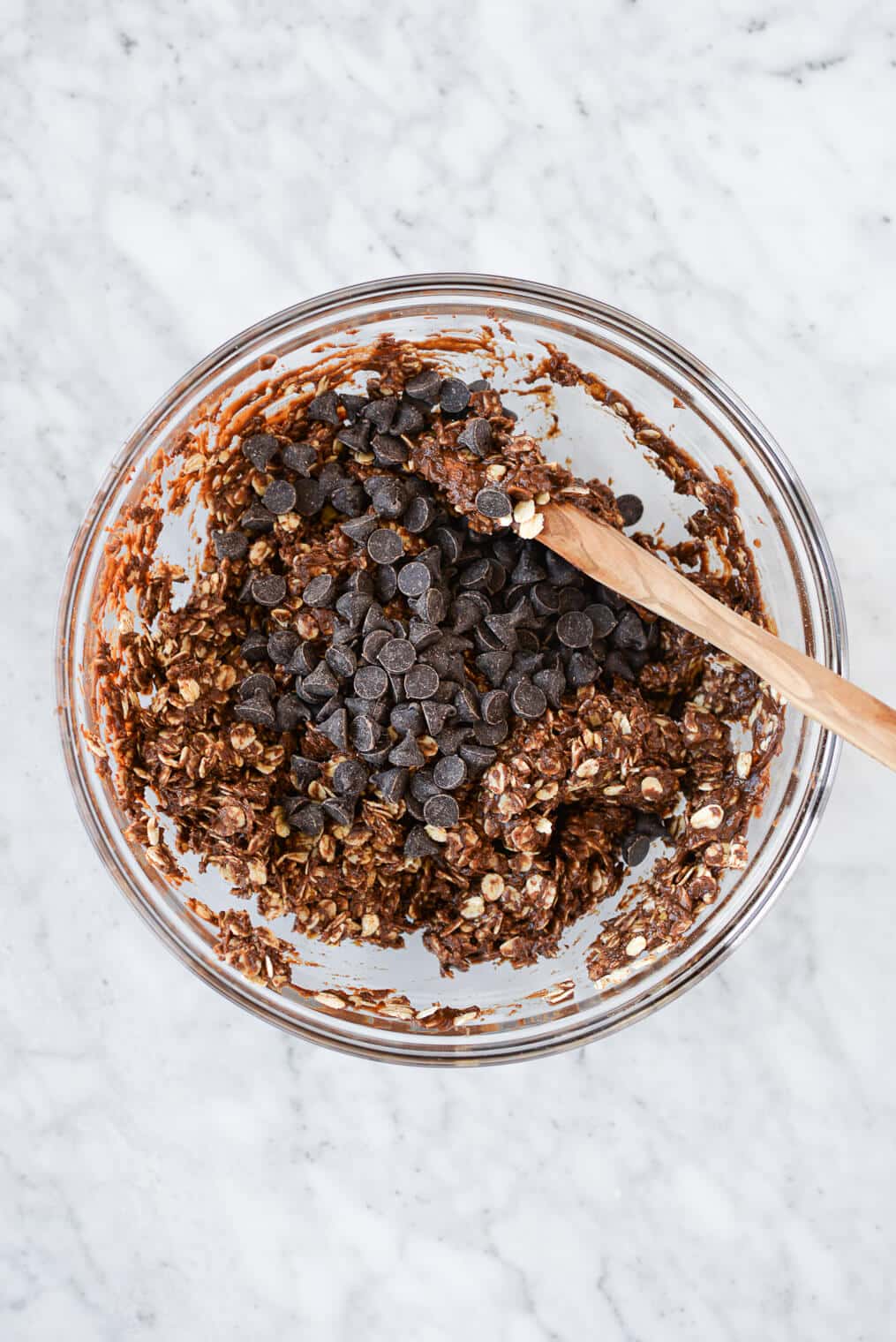 a maple syrup, cocoa powder, coconut oil, and vanilla extract mixture combined with oats and chocolate chips in a large glass bowl with a wooden spoon all on a marble surface