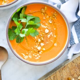 a bowl of bright orange thai carrot soup garnished with fresh cilantro and crushed peanuts sitting on a marble surface