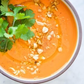 a bowl of bright orange thai carrot soup garnished with fresh cilantro and crushed peanuts sitting on a marble surface