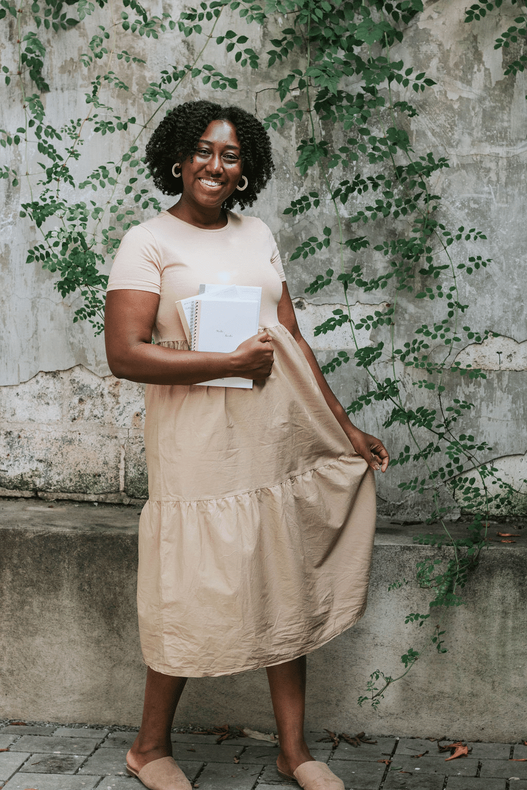 Ashley Brown of Routine & Things standing in front of a wall with greenery growing on it holding notebooks in her hand