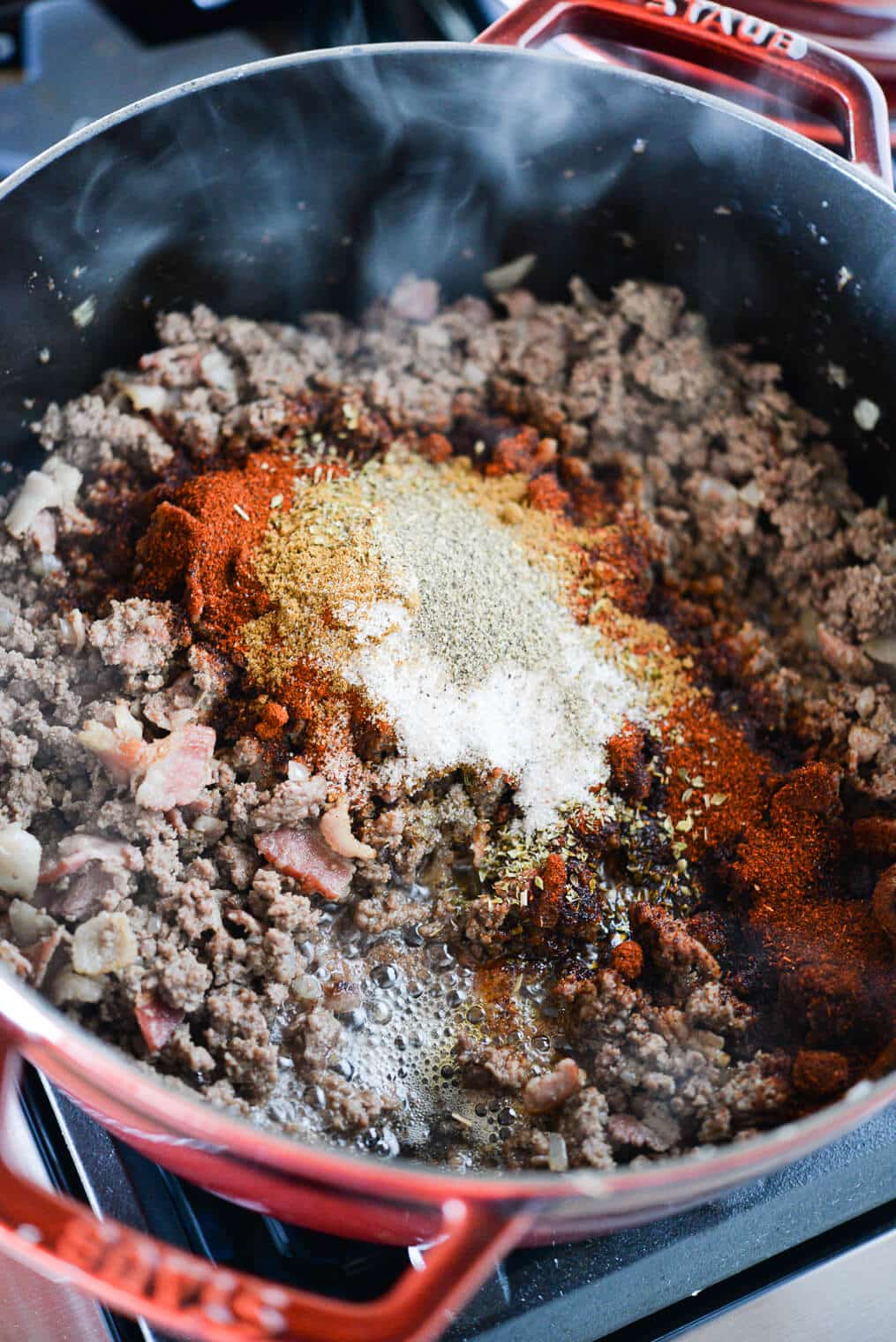 browned ground beef, bacon, onion, and garlic in a large pot with an assortment of spices added on top