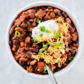 a white bowl filled with beef chili topped with sour cream, green onion, and shredded cheese sitting on a marble surface