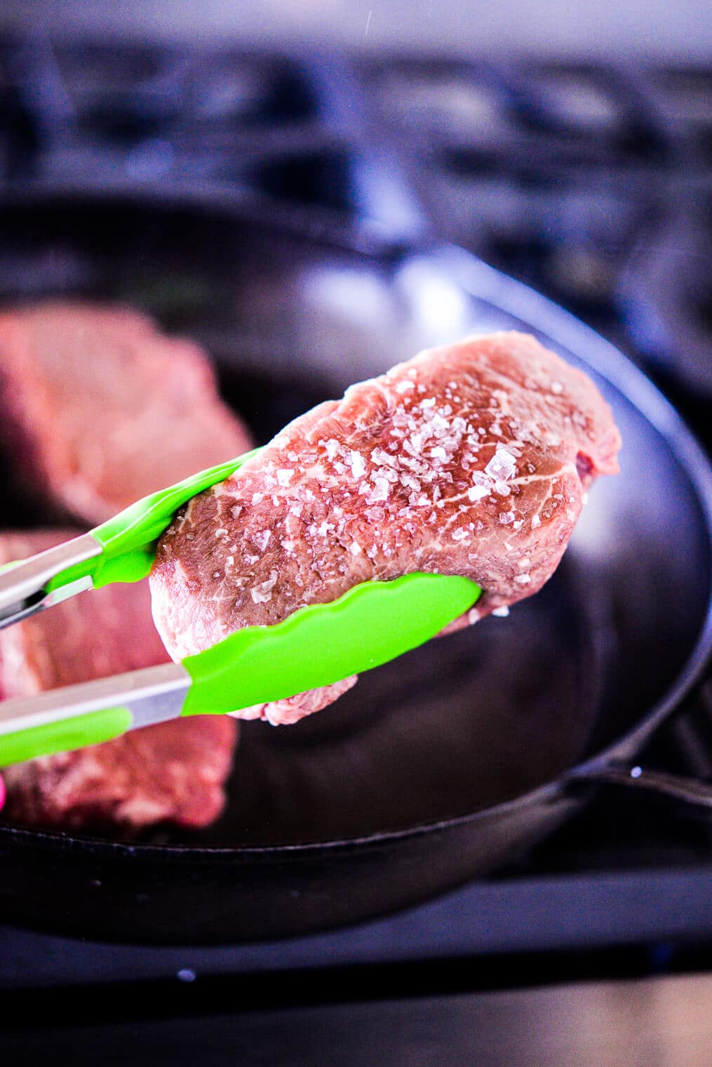 bright green tongs holding a raw, salted filet mignon above a cast iron pan on the stovetop