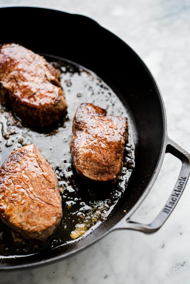 3 cooked filet mignons sitting in a cast iron pan on a marble surface