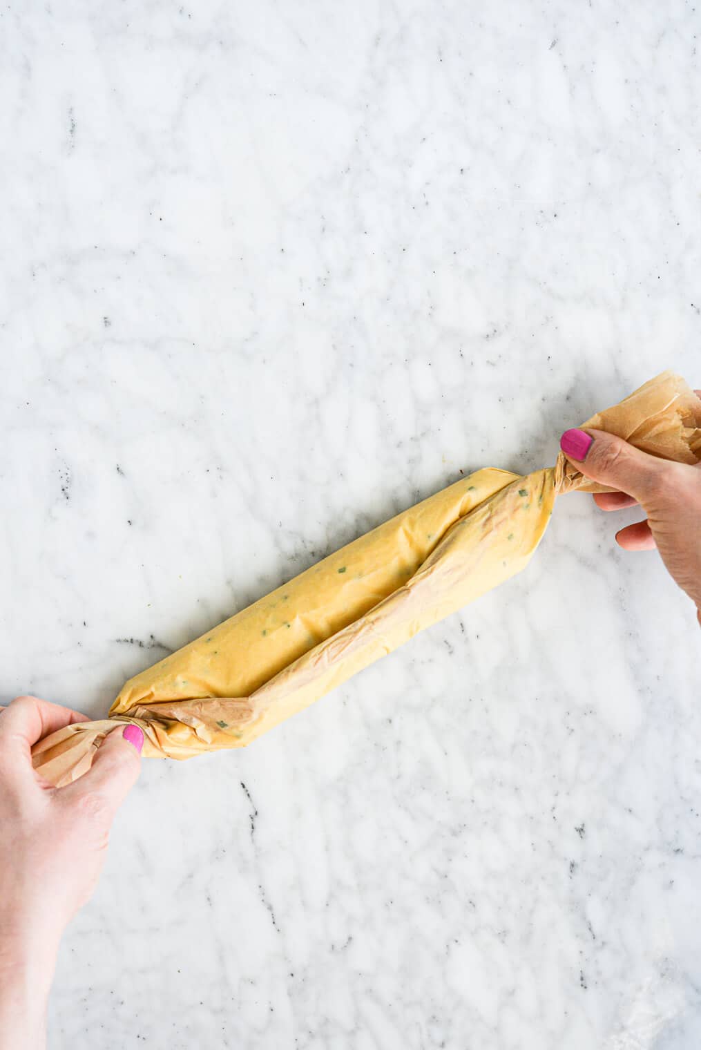 a woman's hands folding parchment paper over a log of compound garlic herb butter. All on a marble surface.