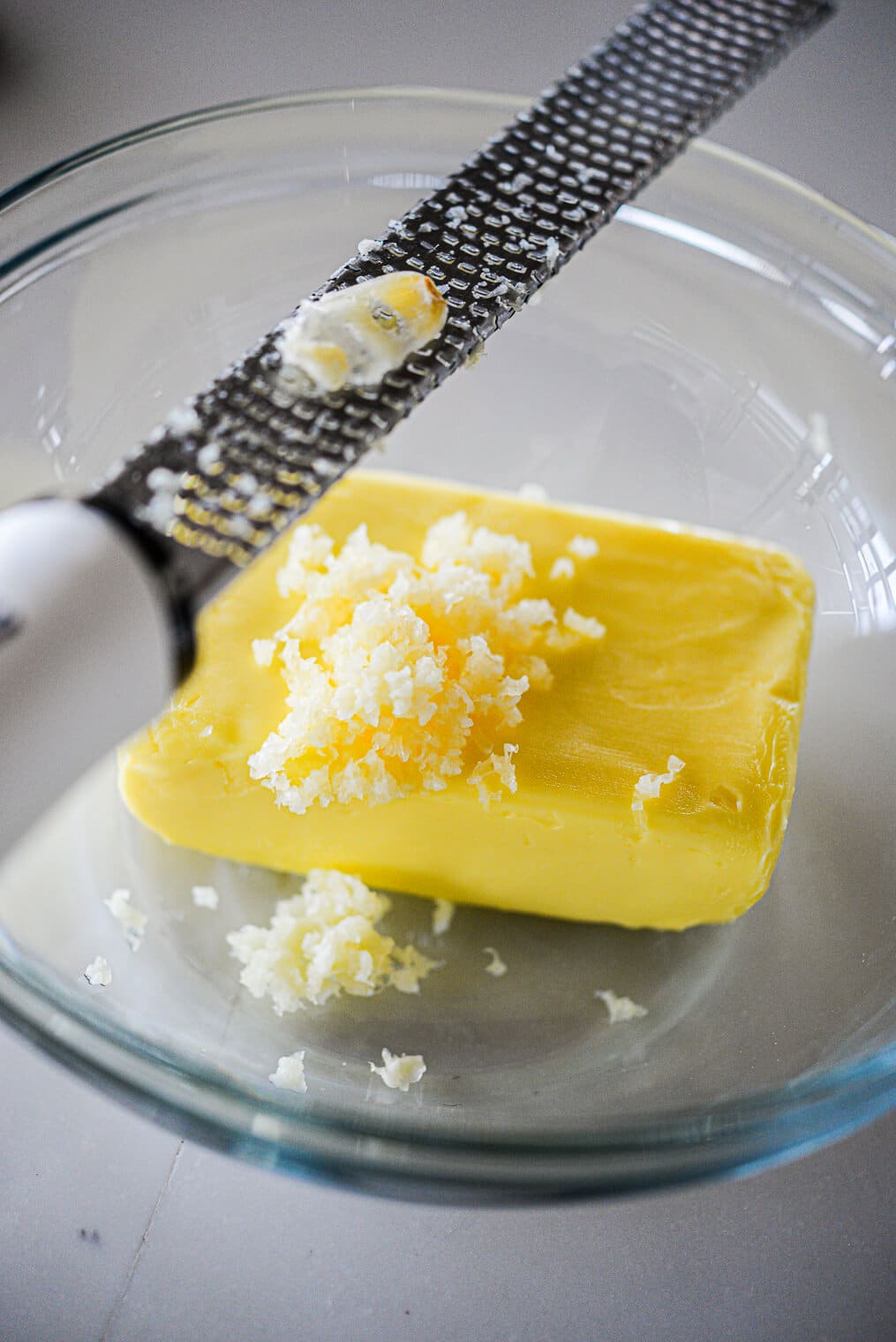 a glass bowl with a block of softened butter in it and a microplane grating garlic into the bowl