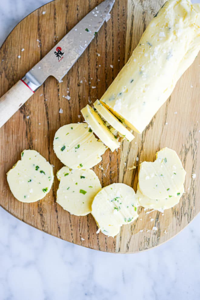 a log of compound garlic and herb butter sliced in 8 pats sitting on a wooden cutting board next to a knife