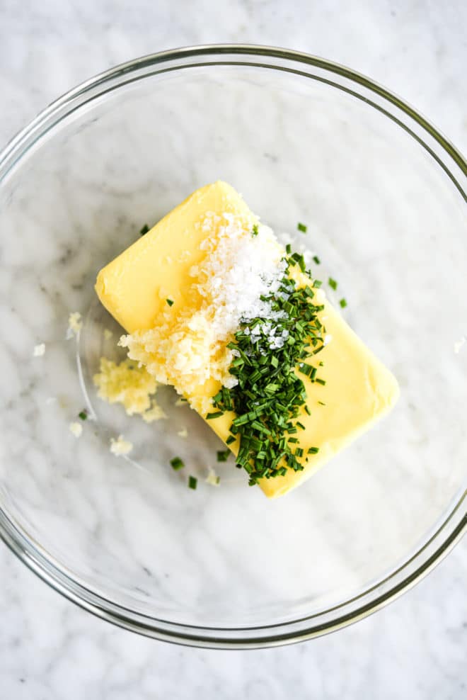 a large block of softened butter with salt, garlic, and chopped herbs sitting on top of it. All on a marble surface.