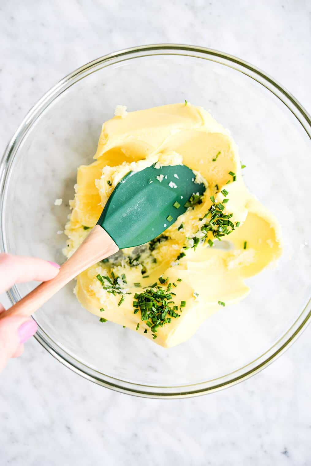 a clear glass bowl with softened butter, herbs, and garlic being mixed together with a spatula. All sitting on a marble surface.