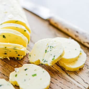 a log of compound garlic and herb butter sliced in 8 pats sitting on a wooden cutting board next to a knife