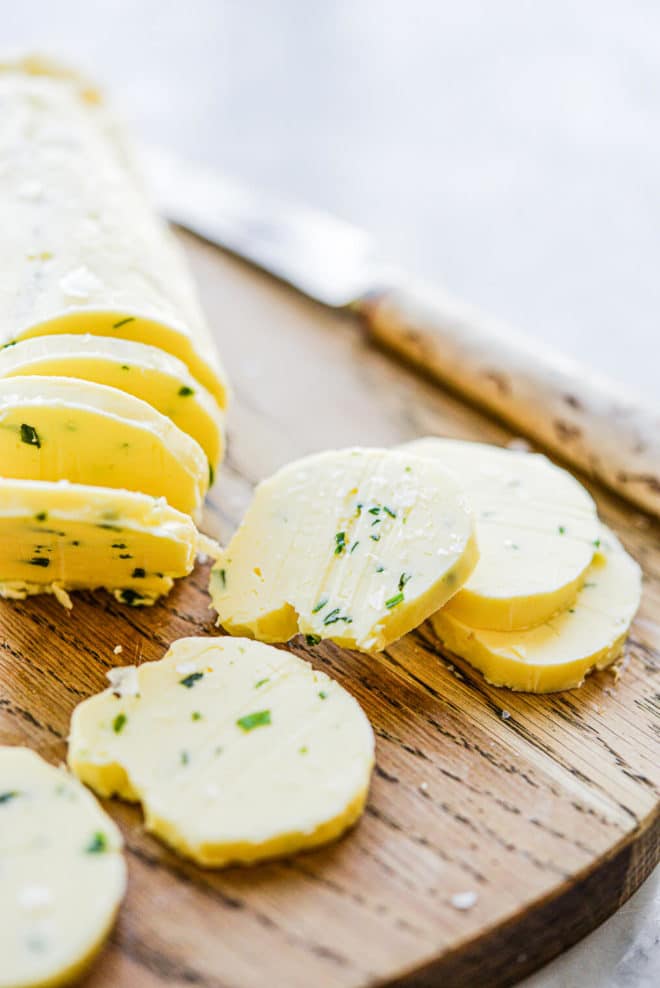 a log of compound garlic and herb butter sliced in 8 pats sitting on a wooden cutting board next to a knife