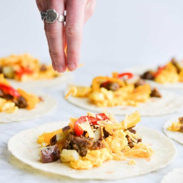 a woman's hand sprinkling shredded cheese over a breakfast taco filled with scrambled eggs, potatoes, bell peppers, and crumbled breakfast sausage