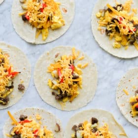 open breakfast tacos filled with scrambled eggs, potatoes, bell peppers, shredded cheese, and crumbed breakfast sausage laying on a marble surface