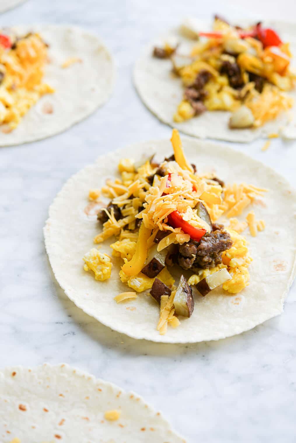 side view of open breakfast tacos filled with scrambled eggs, potatoes, bell peppers, shredded cheese, and crumbed breakfast sausage laying on a marble surface