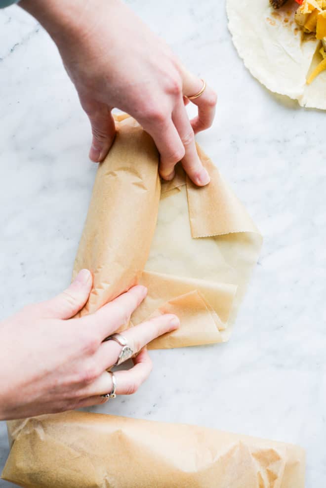 a woman rolling breakfast burritos in parchment paper on a marble surface