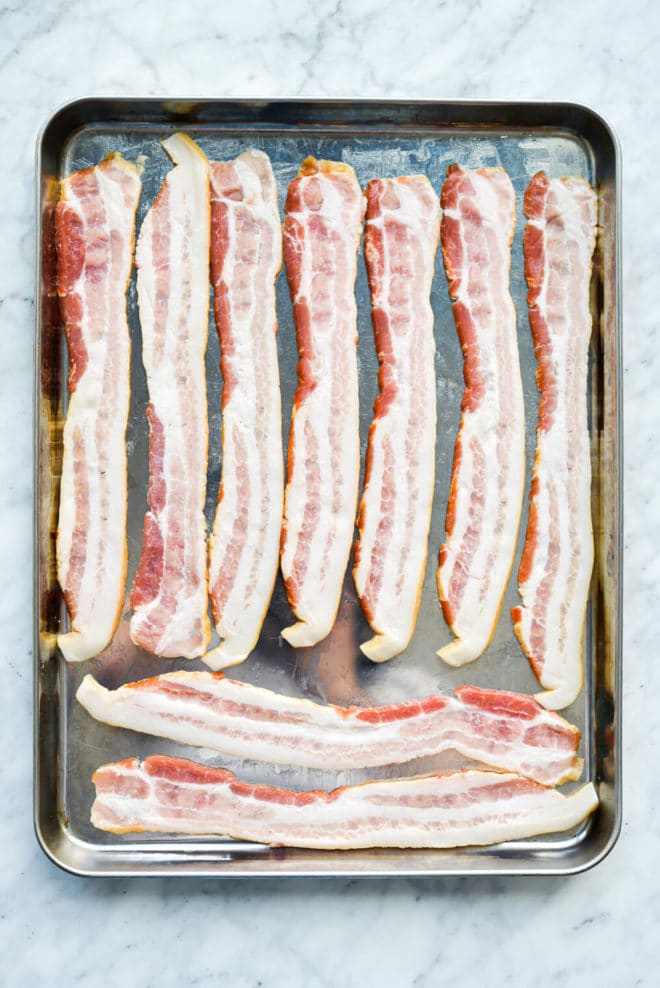 9 slices of raw bacon laying on a sheet pan on a marble surface