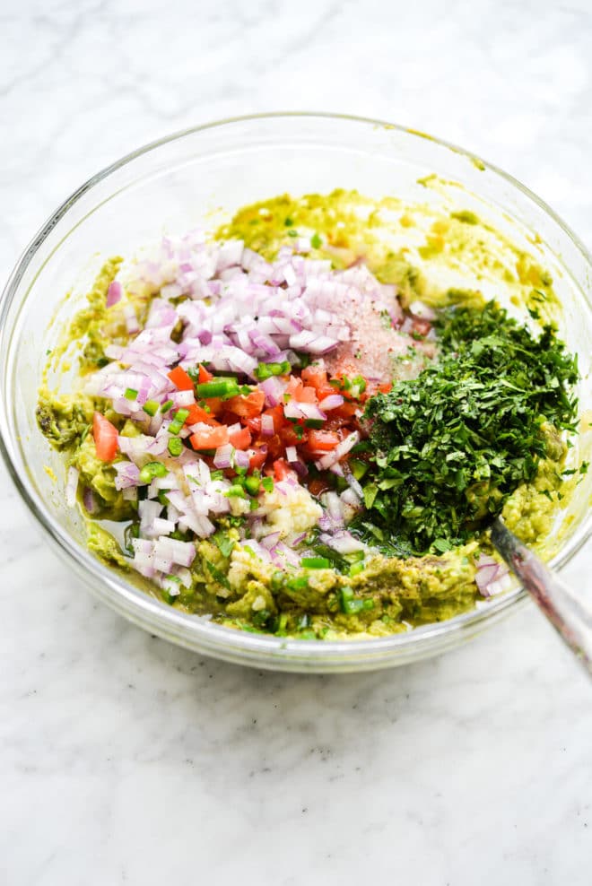 a large glass bowl with mashed avocado, diced red onion, diced tomatoes, diced jalapeno, cilantro, and sea salt on top of it