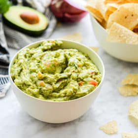 a bowl of guacamole with halved avocados, tortilla chips, tomatoes, red onions, cilantro, and lime halves sitting around it