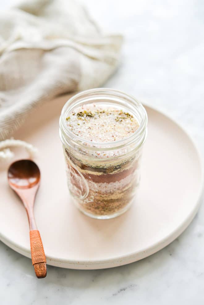 a jar of taco seasoning sitting on a pale pink plate next to a small wooden spoonful of seasoning