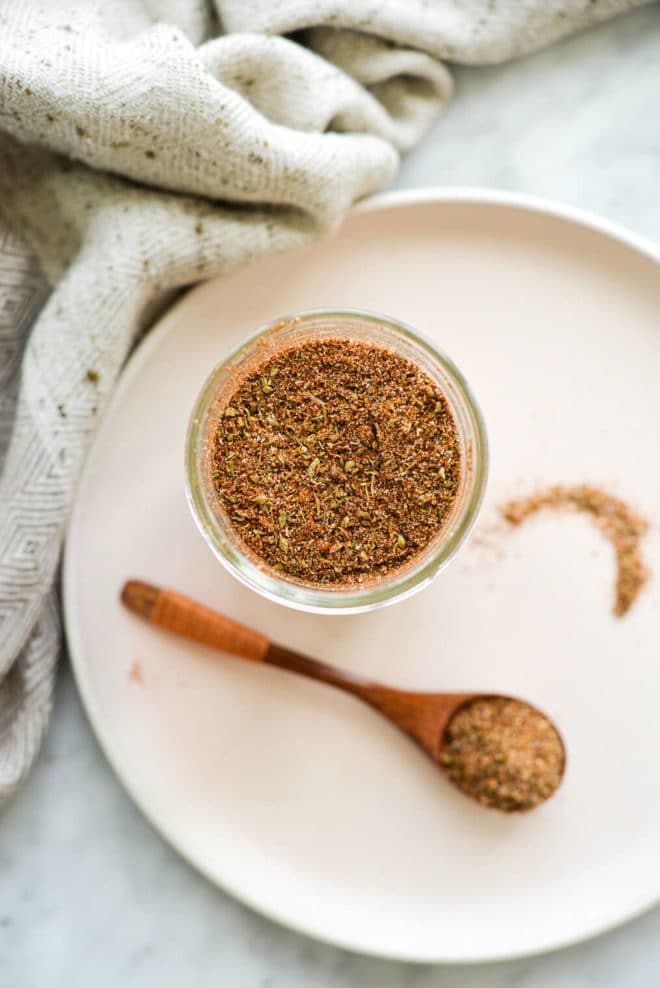 a jar of taco seasoning sitting on a pale pink plate next to a small wooden spoonful of seasoning