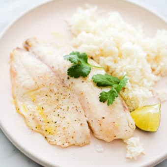 a pale pink plate with a cooked tilapia filet, white rice, a lime wedge, and cilantro sprigs sitting on it, on a marble surface