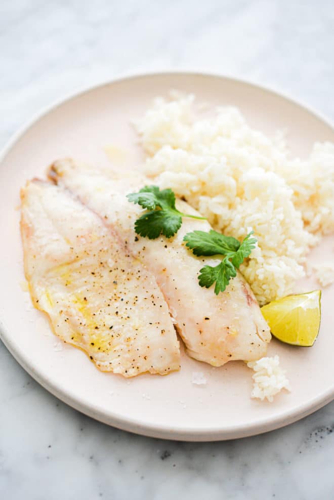 a pale pink plate with a cooked tilapia filet, white rice, a lime wedge, and cilantro sprigs sitting on it, on a marble surface