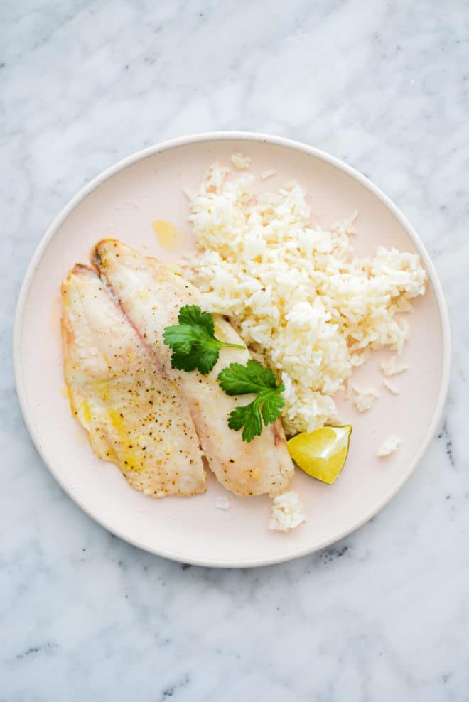 a pale pink plate with a cooked tilapia filet, white rice, a lime wedge, and cilantro sprigs sitting on it, on a marble surface