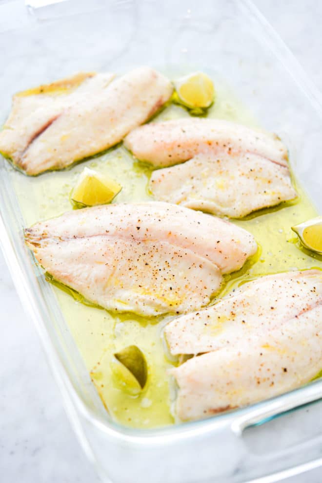 een grote glazen ovenschaal bedekt met olijfolie met 4 gekookte tilapiafilets en 4 limoenpartjes erin