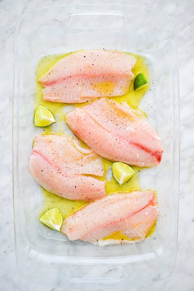 a large glass baking dish coated on the bottom with olive oil with 4 raw tilapia filets and 4 lime wedges sitting in it