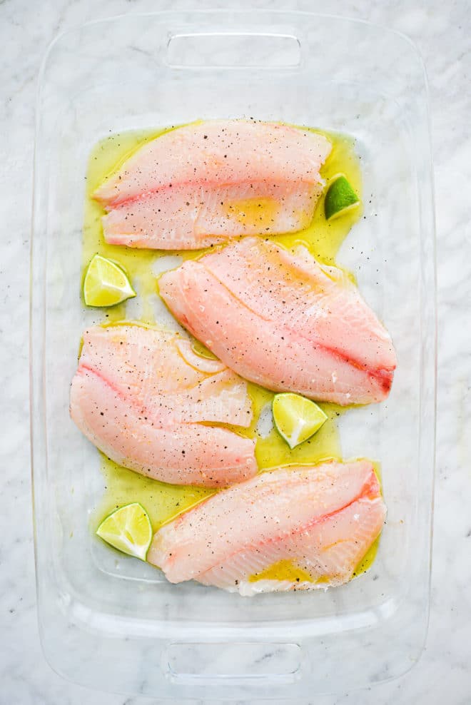 eine große Glasbackform, die unten mit Olivenöl beschichtet ist und in der 4 rohe Tilapia-Filets und 4 Limettenspalten sitzen