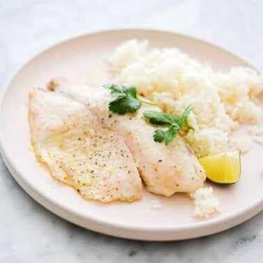 a pale pink plate with a cooked tilapia filet, white rice, a lime wedge, and cilantro sprigs sitting on it, on a marble surface