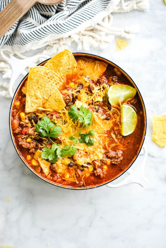a small white pot of taco soup garnished with lime wedges, shredded cheese, cilantro, and tortilla chips