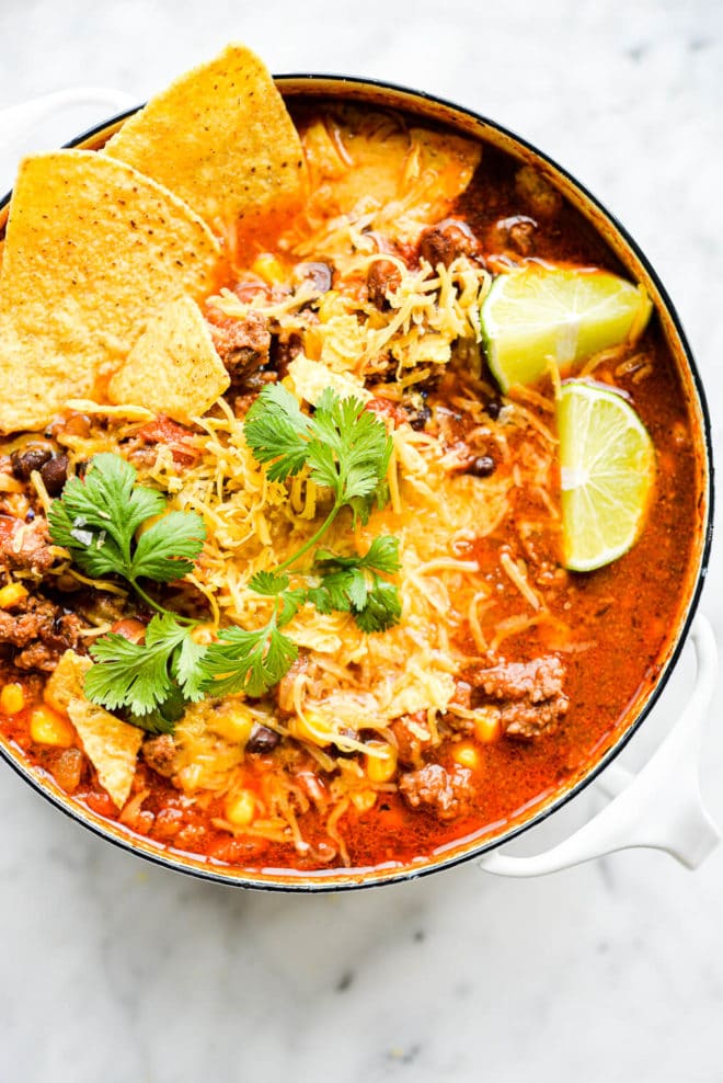 a small white pot of taco soup garnished with lime wedges, shredded cheese, cilantro, and tortilla chips