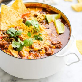 a small white pot of taco soup garnished with lime wedges, shredded cheese, cilantro, and tortilla chips