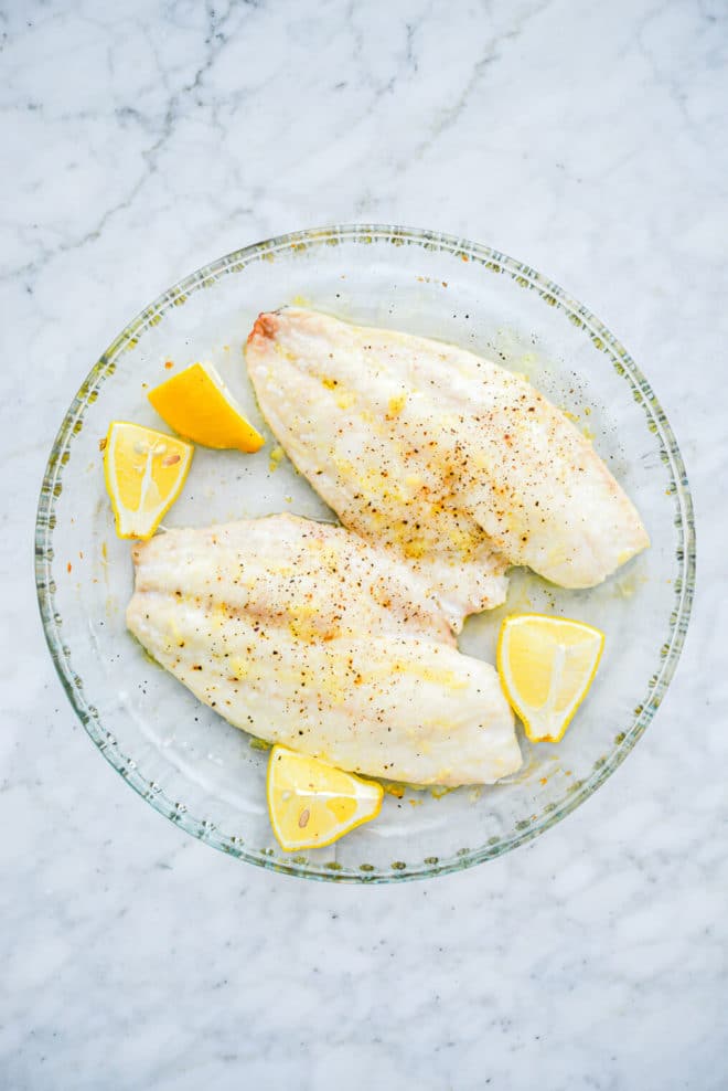a small round glass baking dish with two filets of lightly seasoned red snapper next to 4 lemon wedges