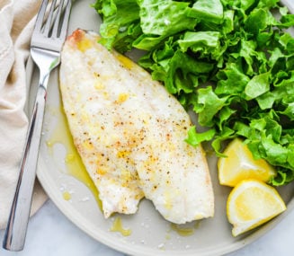 a light gray plate with a filet of red snapper next to a bed of lettuce and lemon wedges