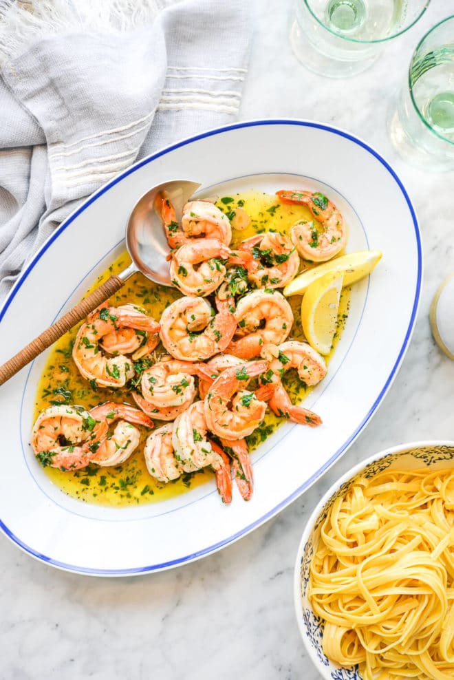 a oblong white dish filled with shrimp scampi sitting on a marble surface next to a bowl of gluten free noodles