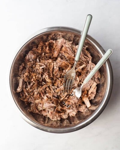 a bowl of shredded pork with seasoning sprinkled on top of it and two forks in the bowl to mix everything together