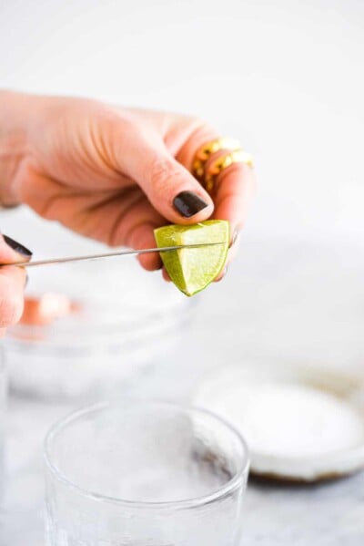 a woman slicing a small lime wedge to use to rub around the rim of a margarita glass before salting it