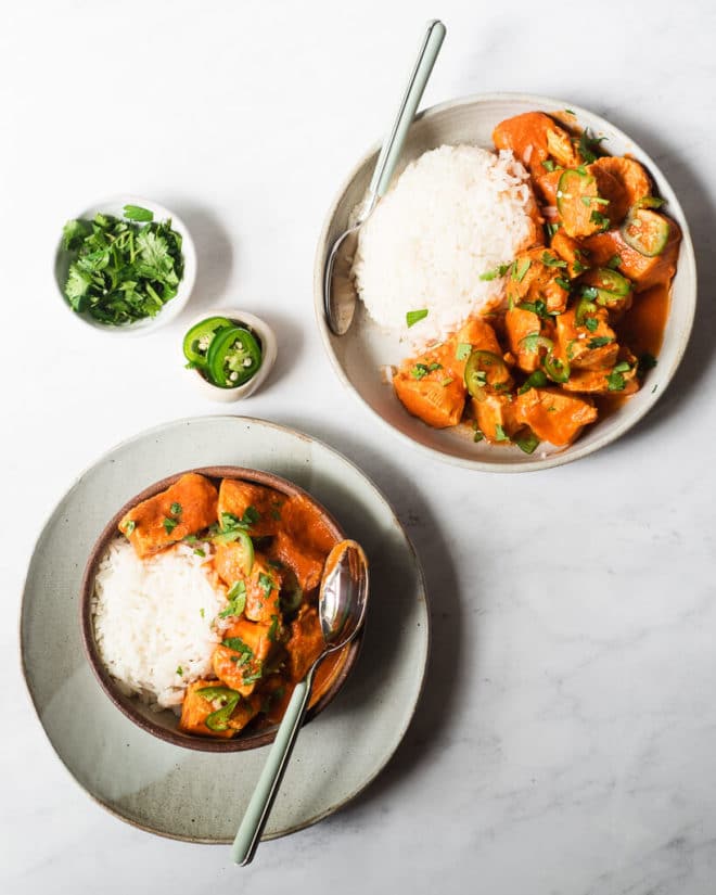 two servings of tikka masala and white rice (one on a plate, and one in a bowl that's sitting on a plate) next to a small bowl of chopped cilantro and a small bowl of sliced raw jalapenos