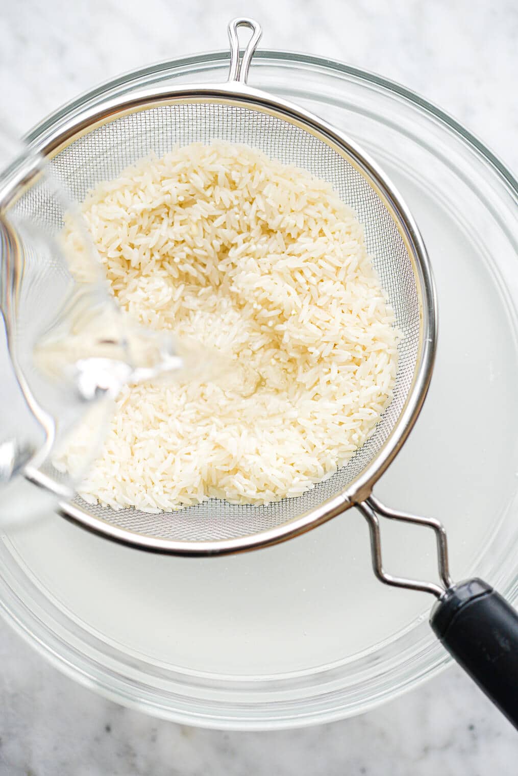 a fine mesh strainer of white rice being rinsed into a large glass bowl