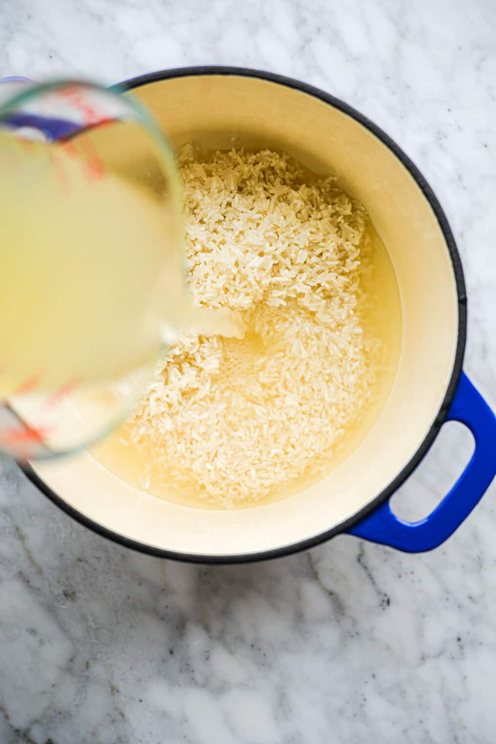 a person pouring chicken broth into a blue and white enameled cast iron pot of uncooked rice